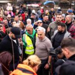 Călin Georgescu în mijlocul unei mulțimi de oameni și jurnaliști, participând la sădirea unor puieți într-o zonă rurală.