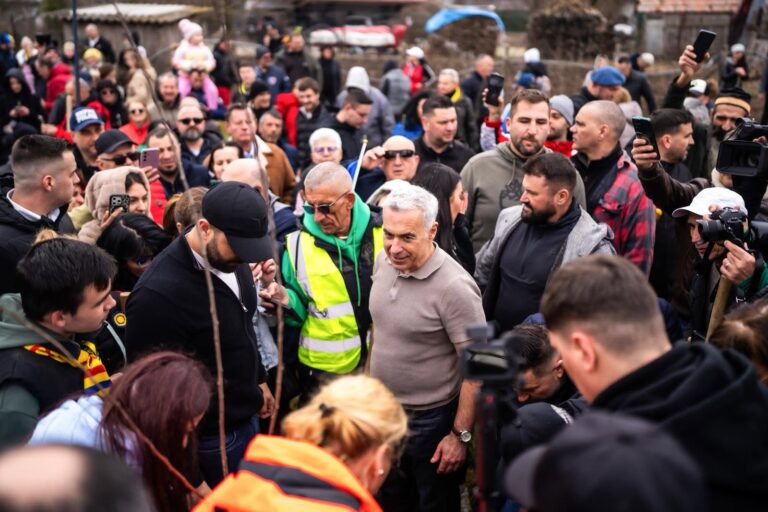 Călin Georgescu în mijlocul unei mulțimi de oameni și jurnaliști, participând la sădirea unor puieți într-o zonă rurală.